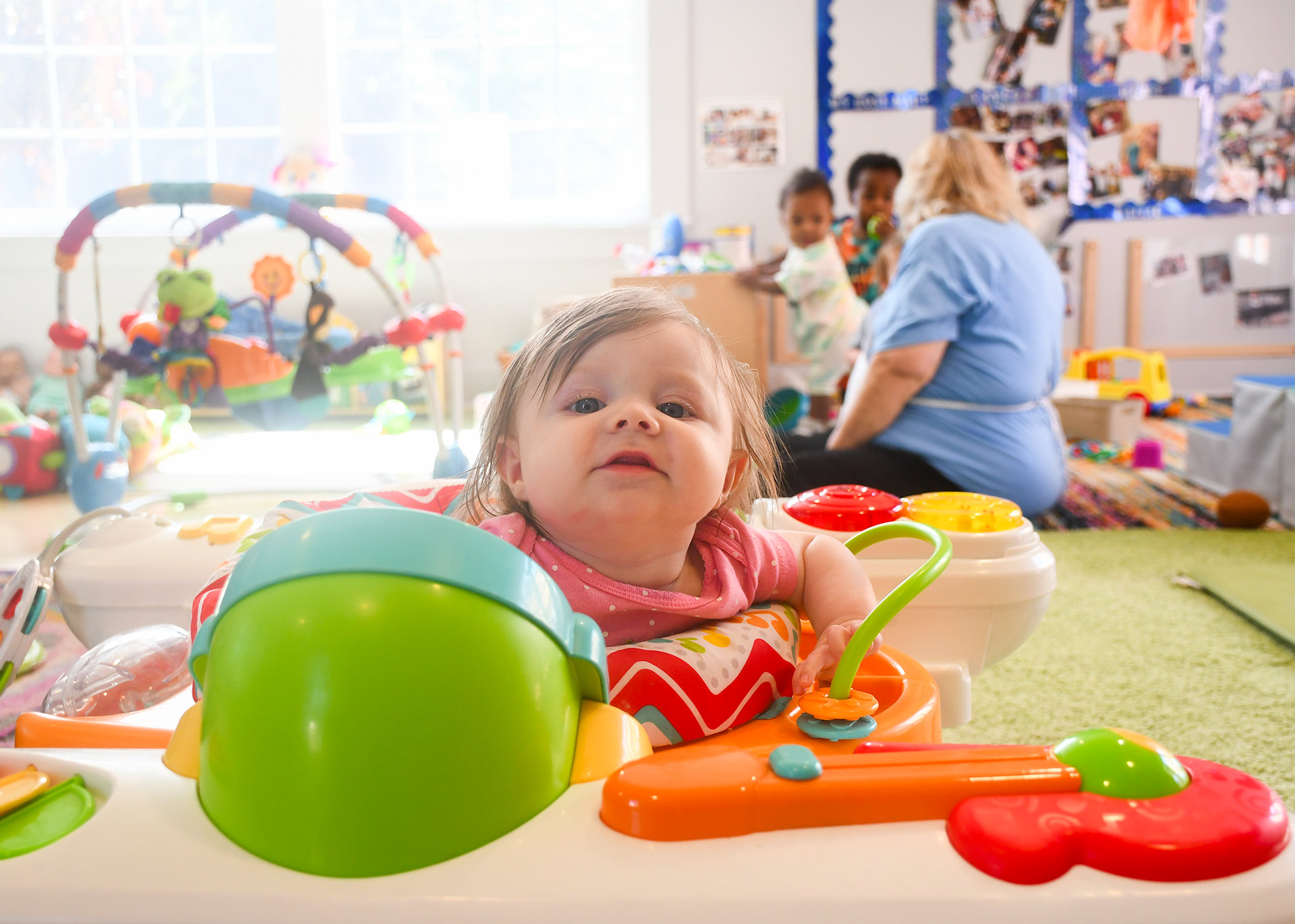 A white baby sits in a bouncy chair in an infant classroom. A teacher and two other babies are behind her.