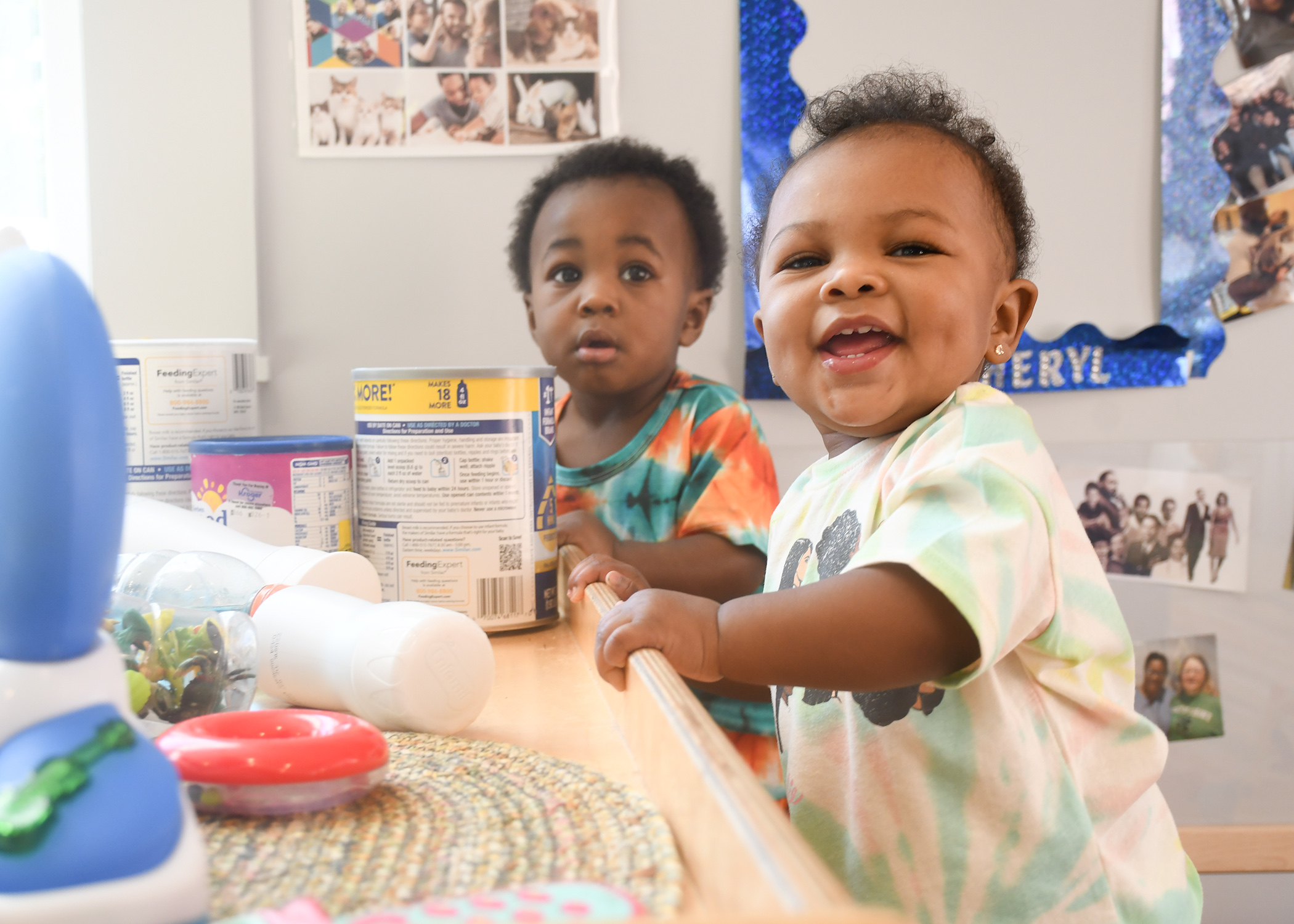 Two African American babies pulling up on a shelf in an infant classroom. Both are looking at the camera and one is smiling.