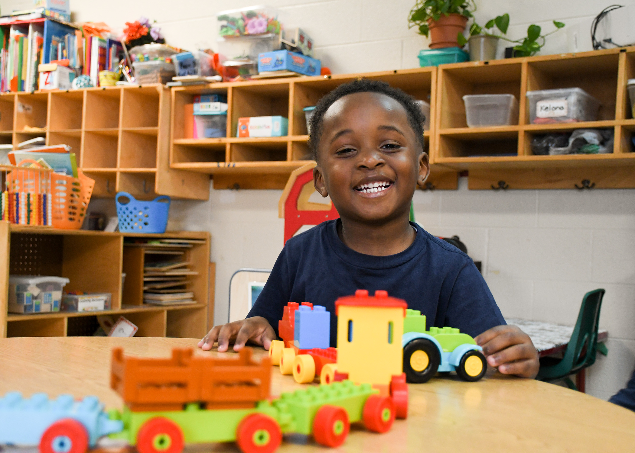 Black boy in a preschool classroom. He's sitting at a table playing with a train and smiling.