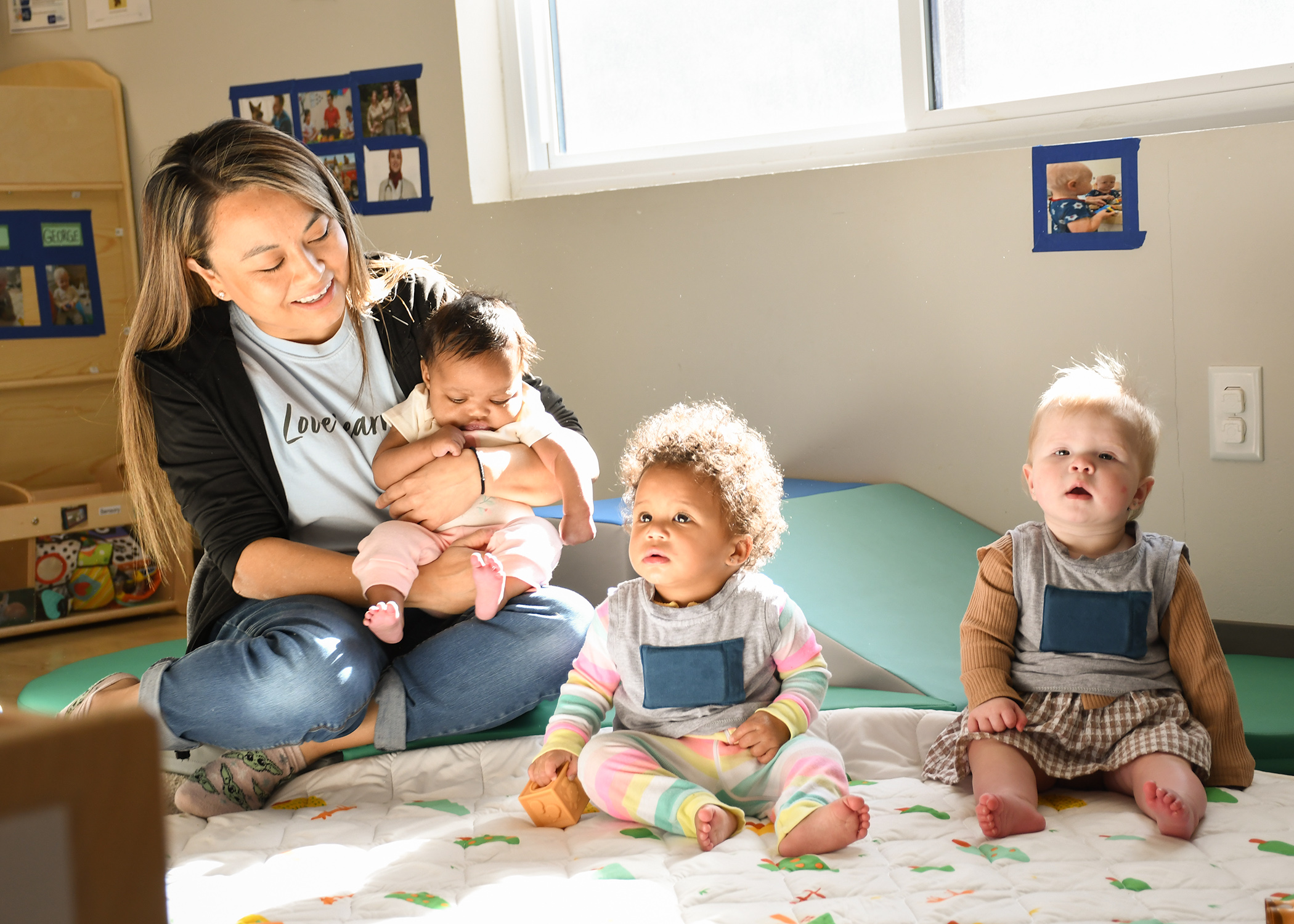 Mixed race child care provider holding an African American child in an infant classroom. A mixed race baby and white baby are sitting on the floor. The room is sunny.