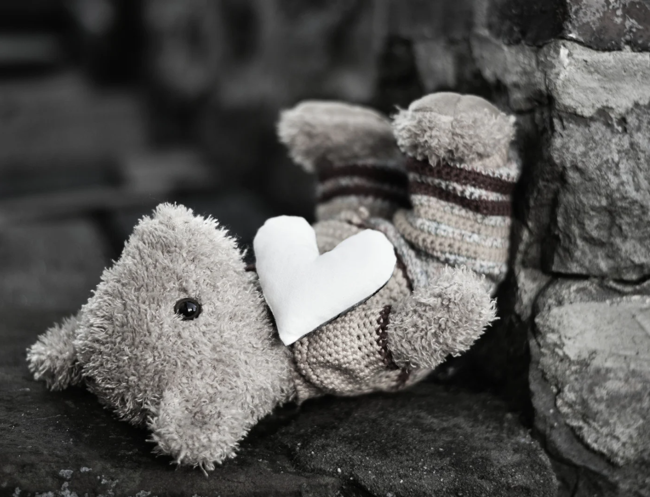 Black and white photo of a teddy bear laying on it's back against a stone wall. A white heart is laying on its chest.